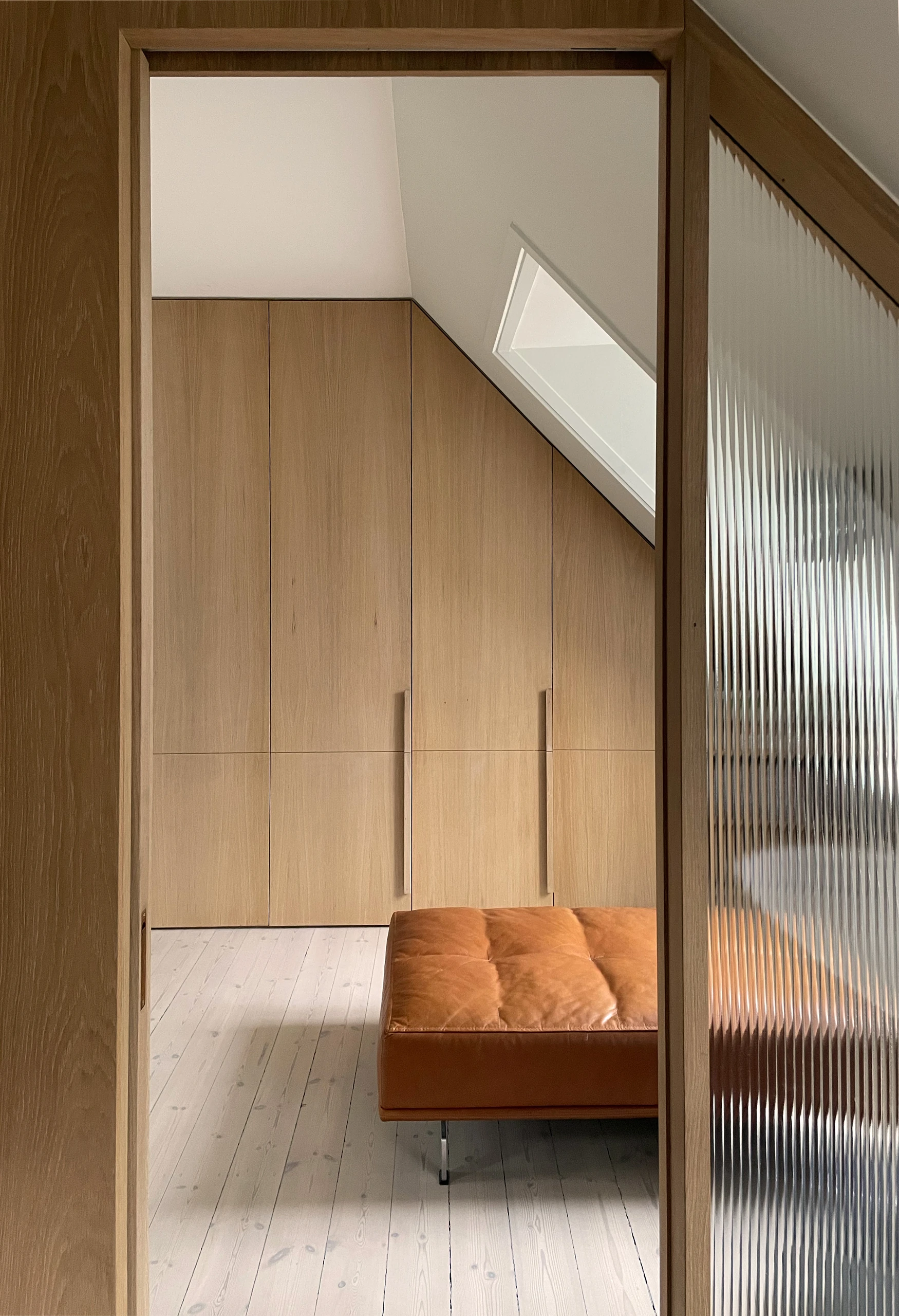 Oak wardrobe wall and tan leather bench seen through a reeded glass door