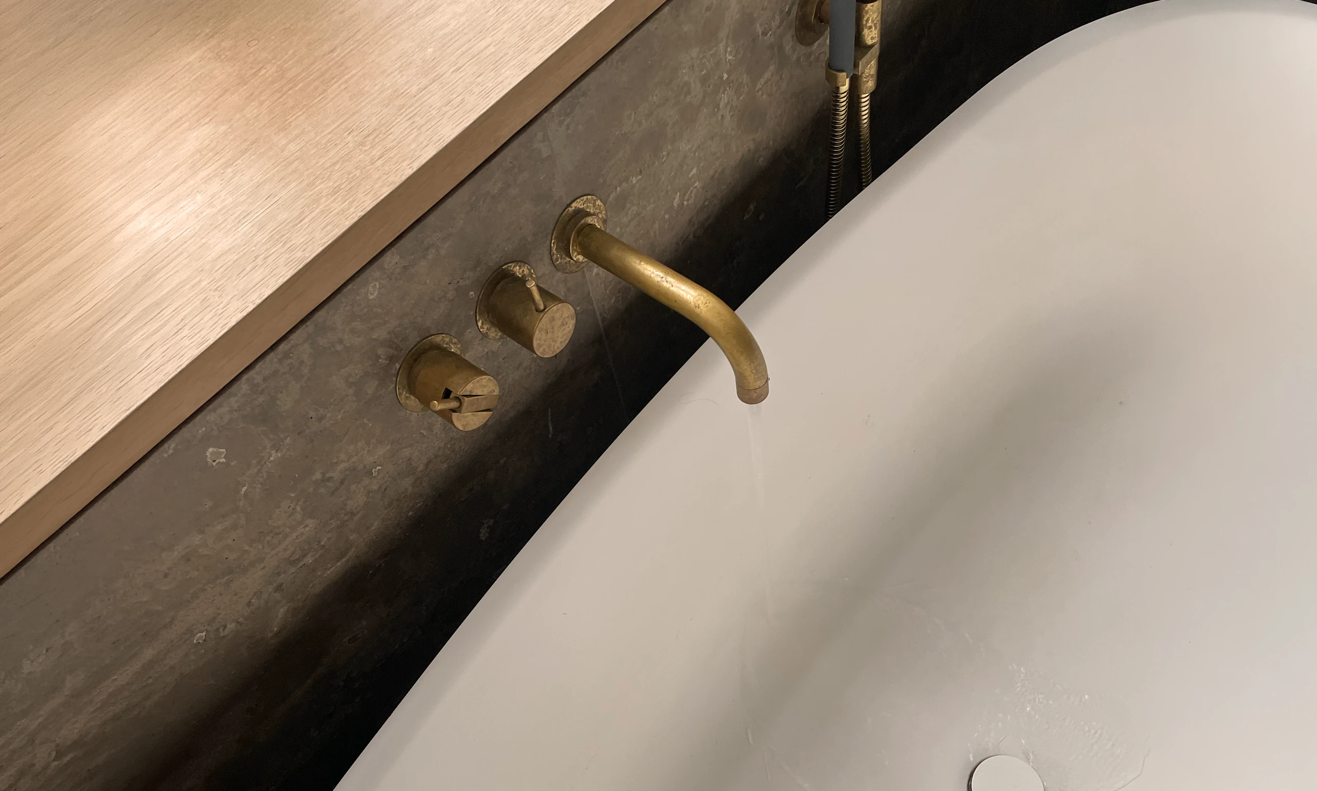 Aged brass tap and mixer set into dark veined stone beside an oak edge and a white bath